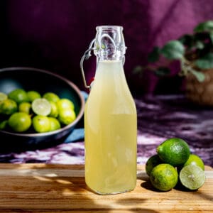 A bottle of key lime simple syrup sits on a wooden cutting board in the sunlight. A small pile of key limes sits on the cutting board next to the bottle of simple syrup.