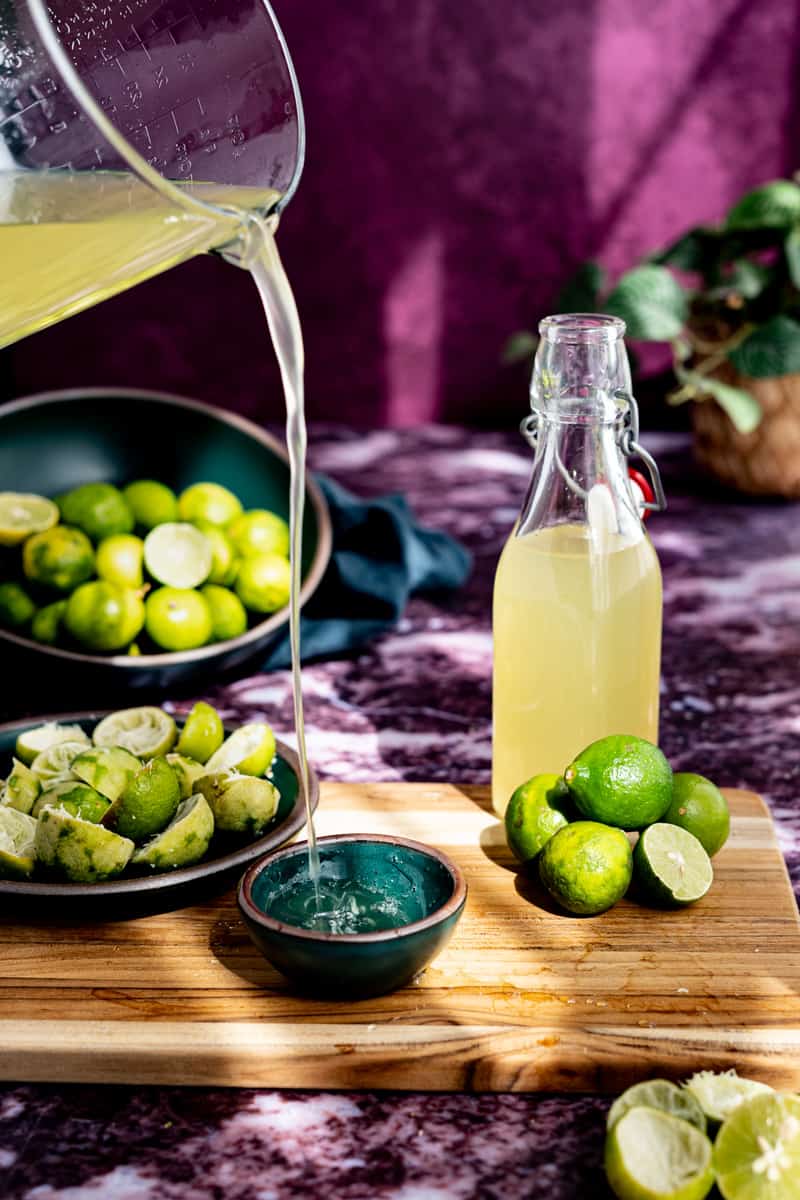 A measuring glass from out of frame is pouring key lime simple syrup into a small teal bowl. A bottle of key lime simple syrup is sitting on a small wooden cutting board next to a pile of key limes.