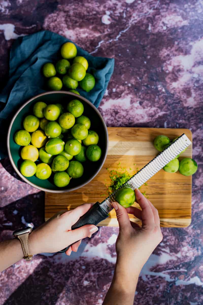 A hand from out of frame is using a microplane zester to zest key limes.