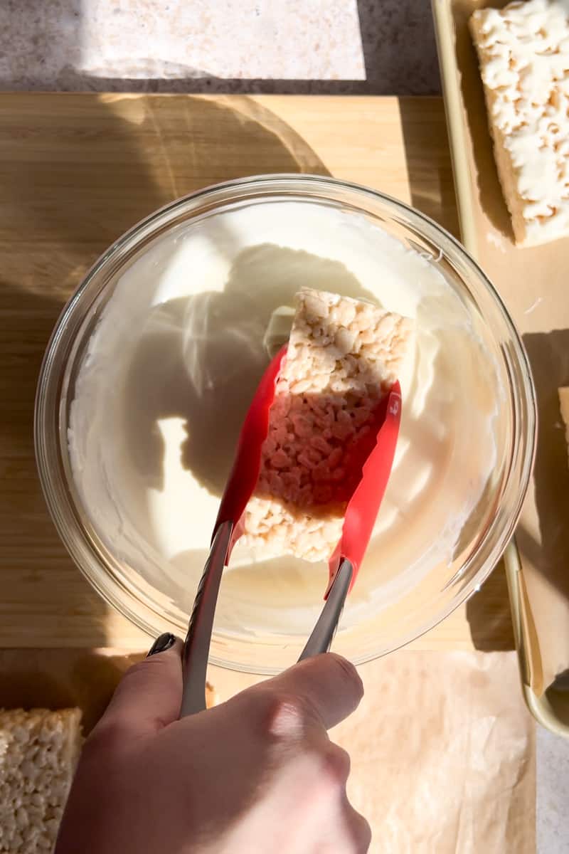 Tongs dipping a rice krispie treat into white chocolate.