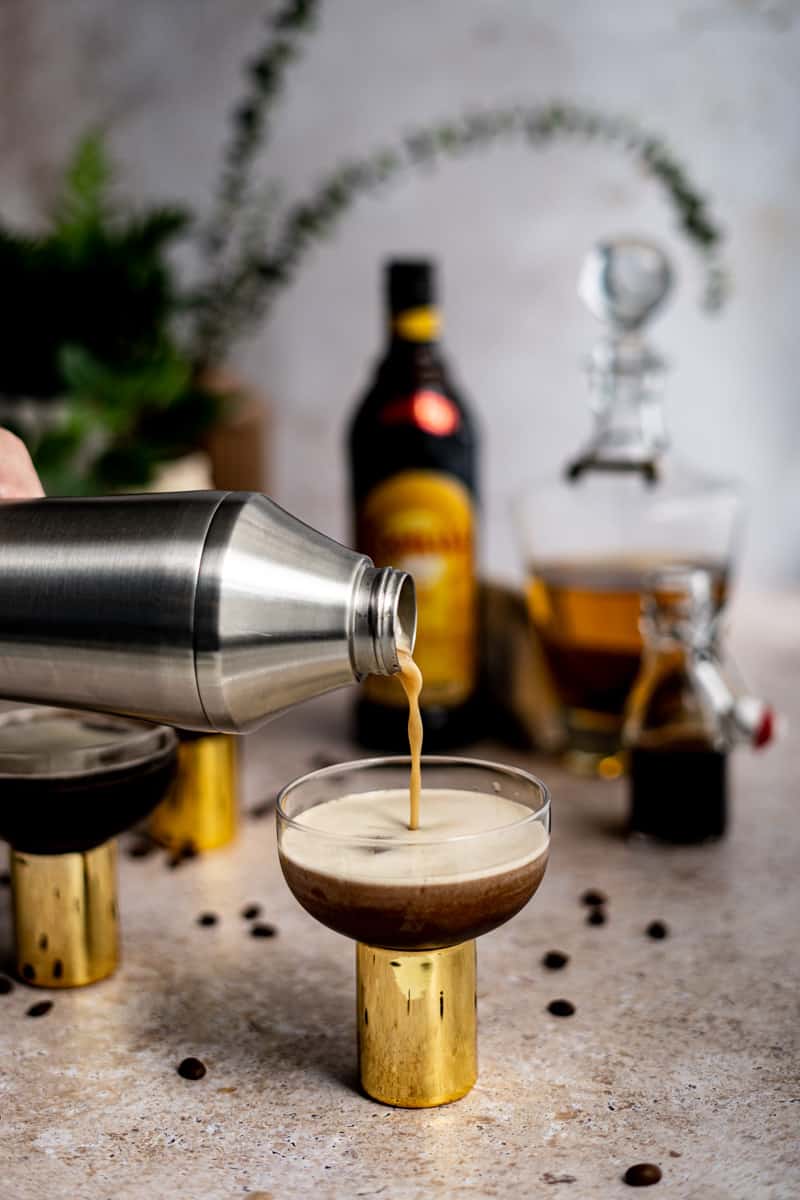 A hand from out of frame is holding a cocktail shaker and pouring a rum espresso martini into a coupe glass.