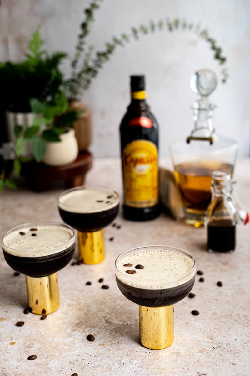 Feature image for a rum espresso martini. Three rum espresso martinis sit on a countertop that is scattered with espresso beans. Ingredients used to make the martinis sits in the background.