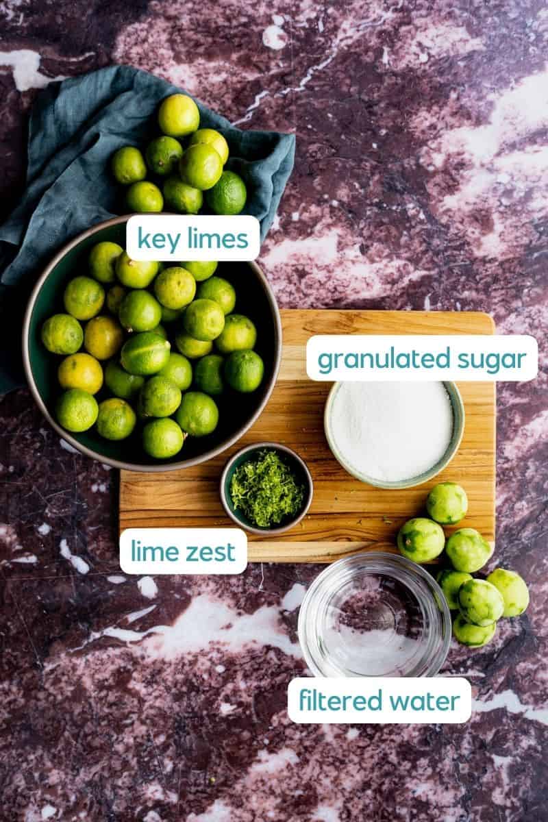 The ingredients used to make key lime simple syrup sit in small bowls on a wooden cutting board against a red marble countertop.