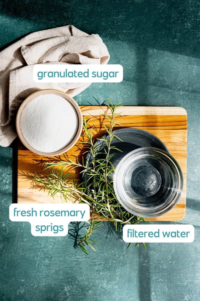Ingredients used to make rosemary simple syrup sit on a small cutting board on a green countertop. The photo is labeled with the titles of the ingredients including: granulated sugar, filtered water, and fresh sprigs of rosemary.