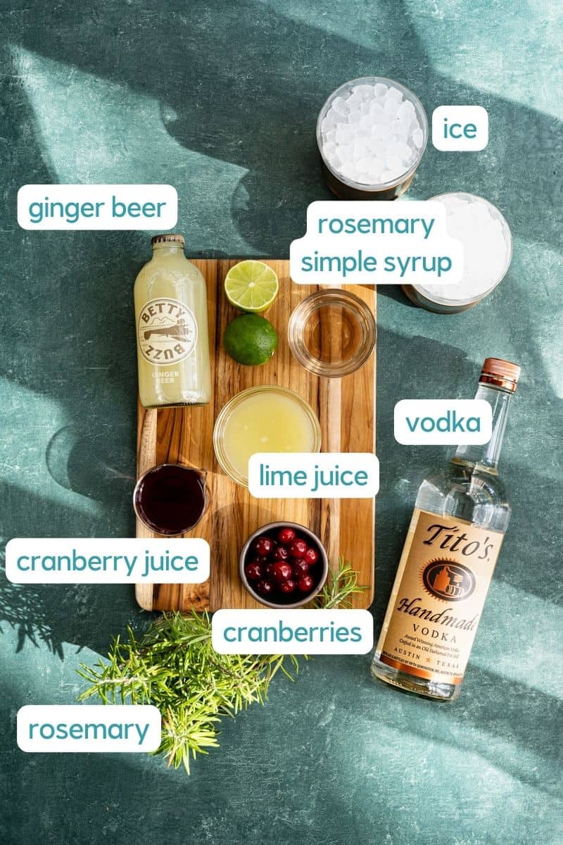 Ingredients used to make a Yule Mule cocktail sit on a wooden cutting board on a green countertop. The photo is labeled with names of each ingredient.