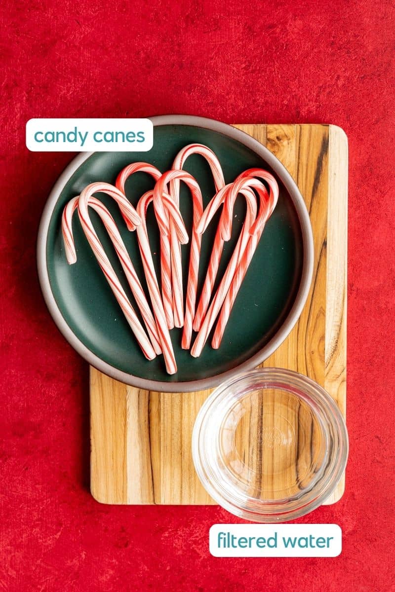 The ingredients used to make a candy cane simple syrup sit on a wooden cutting board on a red countertop. The ingredients are labeled.