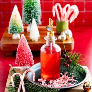 A bottle of candy cane simple syrup sits on a dish on a countertop surrounded by Christmas decor and crushed candy cane pieces.