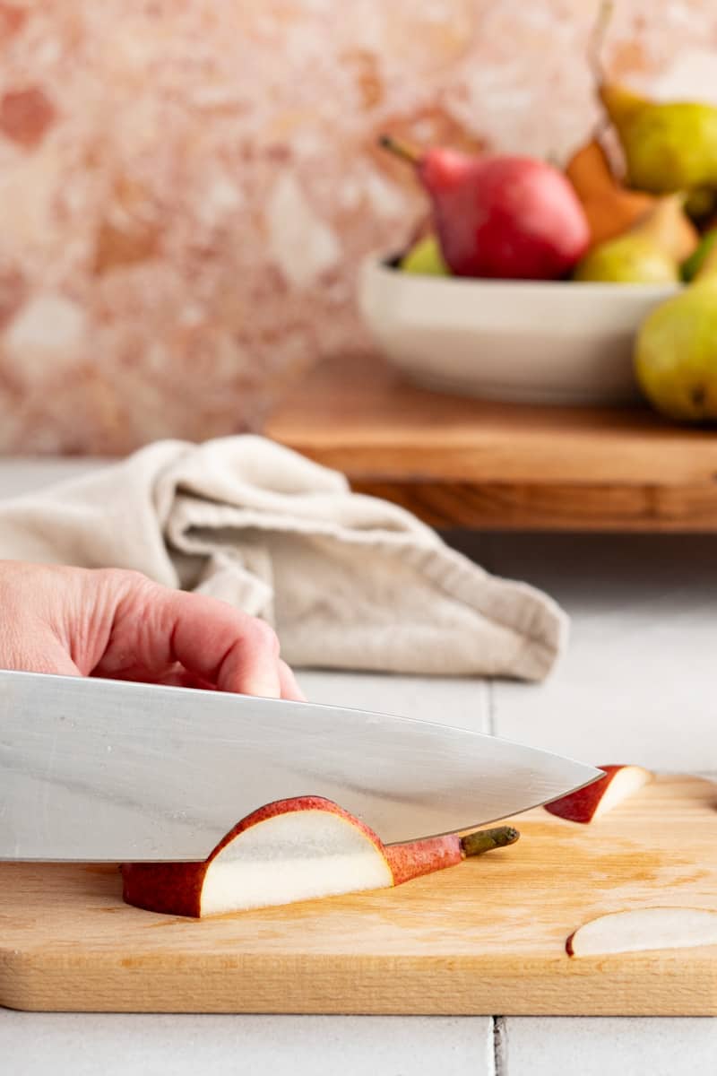 A knife being held out of frame is thinly slicing pears to use as a garnish in a autumn rum punch cocktail.