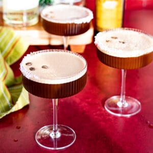 Three peppermint espresso martinis sit on a red countertop with ingredients used to make the drink in the background.
