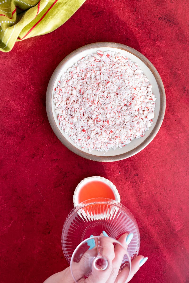 A hand from out of frame lightly presses a cocktail coupe into a small dish of candy cane simple syrup to rim the glass.