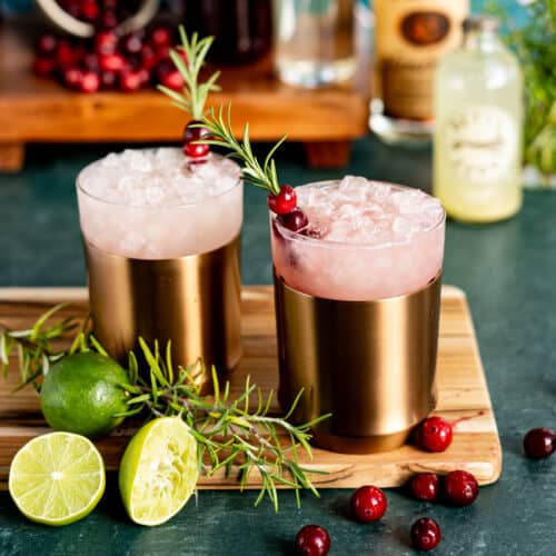 Two Yule Mule cocktails sit on a wooden cutting board with the ingredients used to make them in the background.