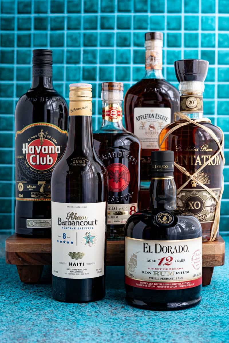 Six bottles of aged rum are sitting against a teal countertop. The brands include Havana Club, Rhum Barbancourt, Appleton, Bacardi, Plantation, and El Dorado.