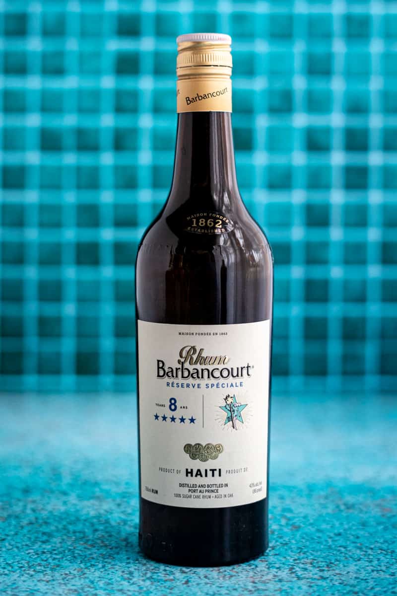 A bottle of Rhum Barbancourt 8 Year old aged Haitian Rum sits on a teal countertop.