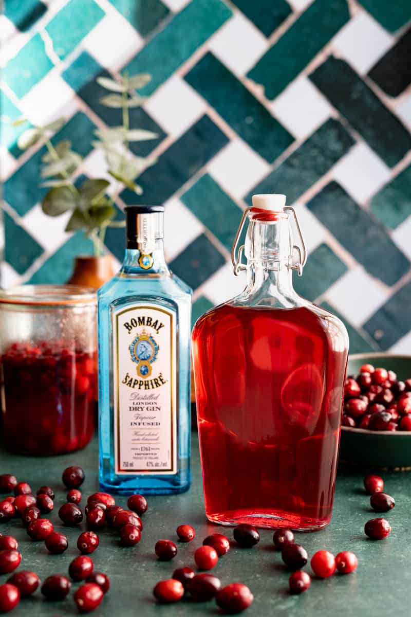 A bottle of homemade cranberry infused gin sits on a green countertop with cranberries scattered around it. There is a jar of the cranberries infusing in gin in the background, a bottle of gin, and a bowl of cranberries in the background.