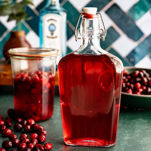 A bottle of homemade cranberry infused gin sits on a green countertop with cranberries scattered around it. There is a jar of the cranberries infusing in gin in the background and a bowl of cranberries in the background.