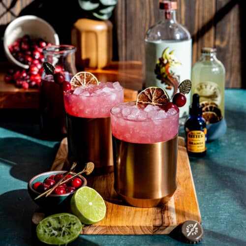 Feature image of a zero-proof cranberry mule beverage. Two copper mule glasses sit on a wooden cutting board, garnished with lime and cranberries. There are limes in the foreground and ingredients used to make the mules in the background.