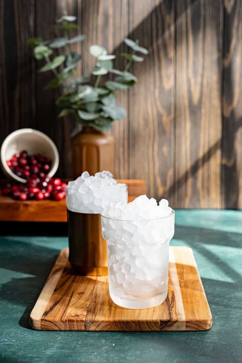 Two cocktail glasses filled with ice sit on a cutting board. One has a copper sleeve on it, the other is just glass so you can see the ingredients.