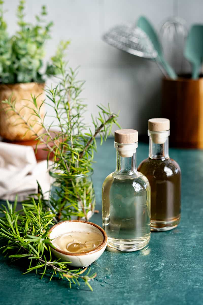 Two bottles of rosemary simple syrup sit on a green countertop with rosemary on the countertop as well. A small bowl has syrup being poured into it. One bottle of syrup has been infused longer and with more rosemary, so it is darker than the other.
