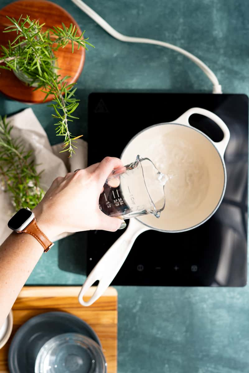 A hand from out of frame is pouring water into a saucepan and heating it over medium heat.
