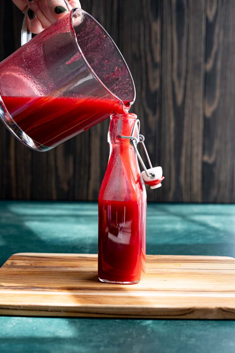 Pouring the finished spiced cranberry syrup into a glass storage bottle.