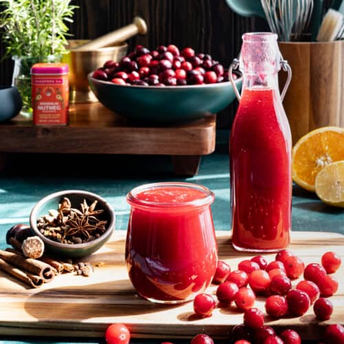 Spiced cranberry syrup sits in a small glass jar. A glass bottle of spiced cranberry syrup sits next to the small jar, and cranberries are scattered in the foreground.