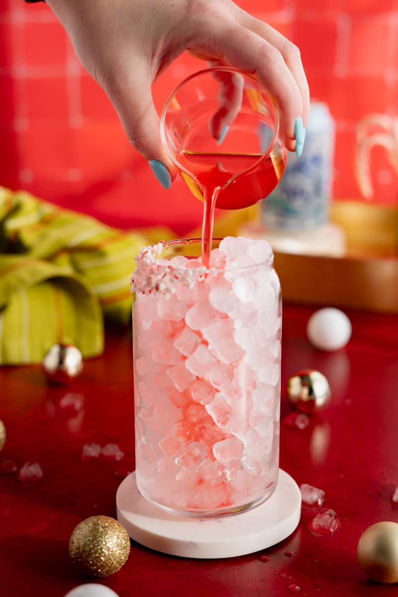 A glass filled with ice has candy cane simple syrup being poured into it.