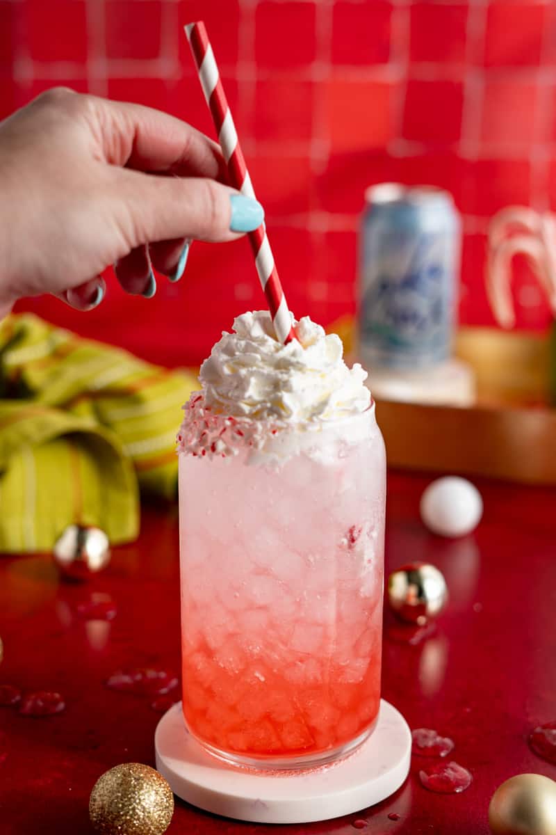 A hand from out of frame puts a red and white striped straw into a candy cane soda.