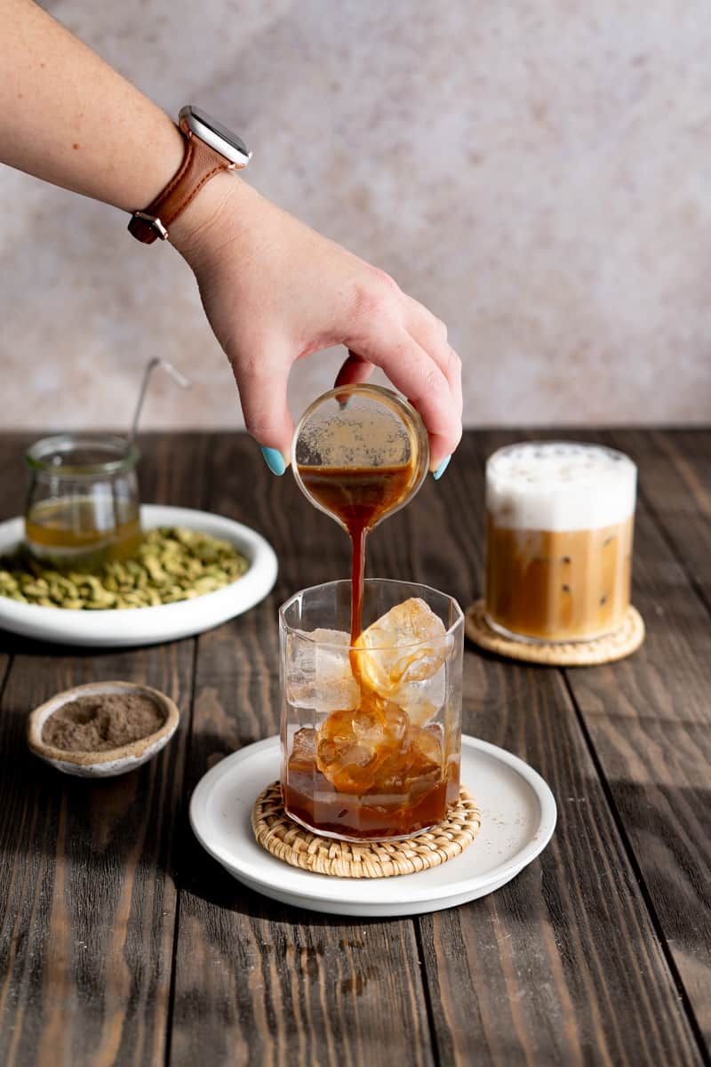 A hand from out of frame pours slightly cooled espresso into a glass filled with ice cubes.