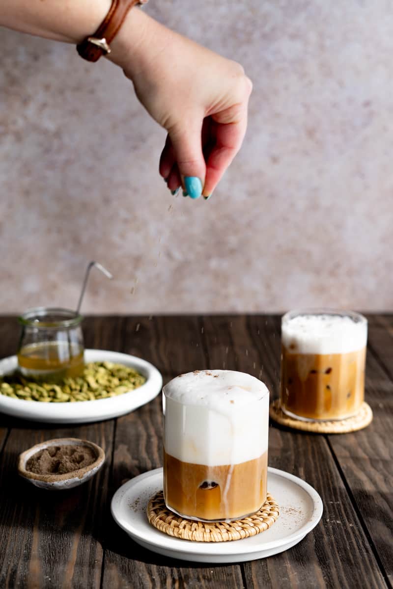 A hand from out of frame sprinkles ground cardamom onto an iced cardamom latte.