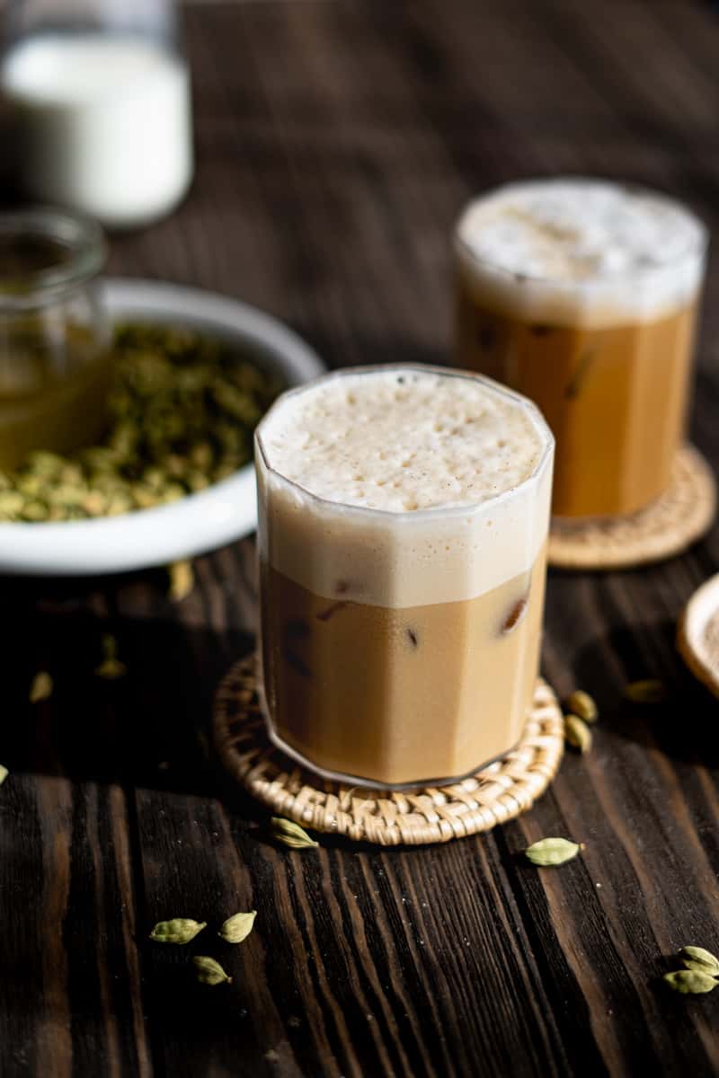 Two glasses of iced cardamom lattes sit on a wooden countertop. There are cardamom pods sprinkled on the countertop and a small dish of ground cardamom in the background.