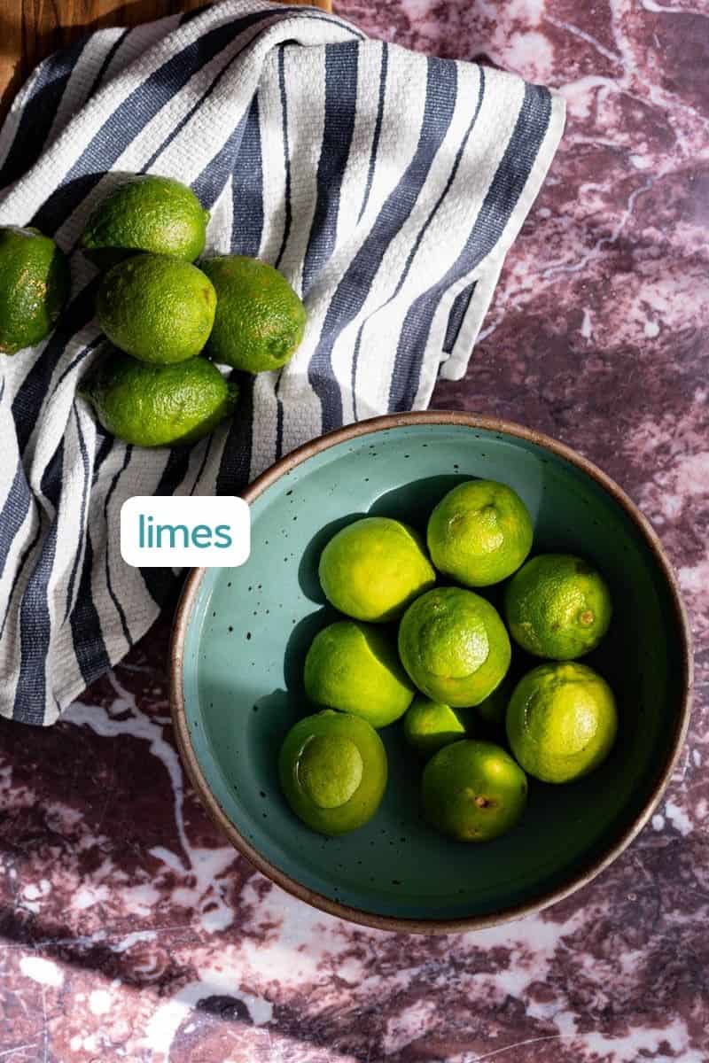 A labeled ingredient shot for a dehydrated lime wheel recipe. A bowl of limes sits on a red marble countertop with a hand towel drying limes to the left.