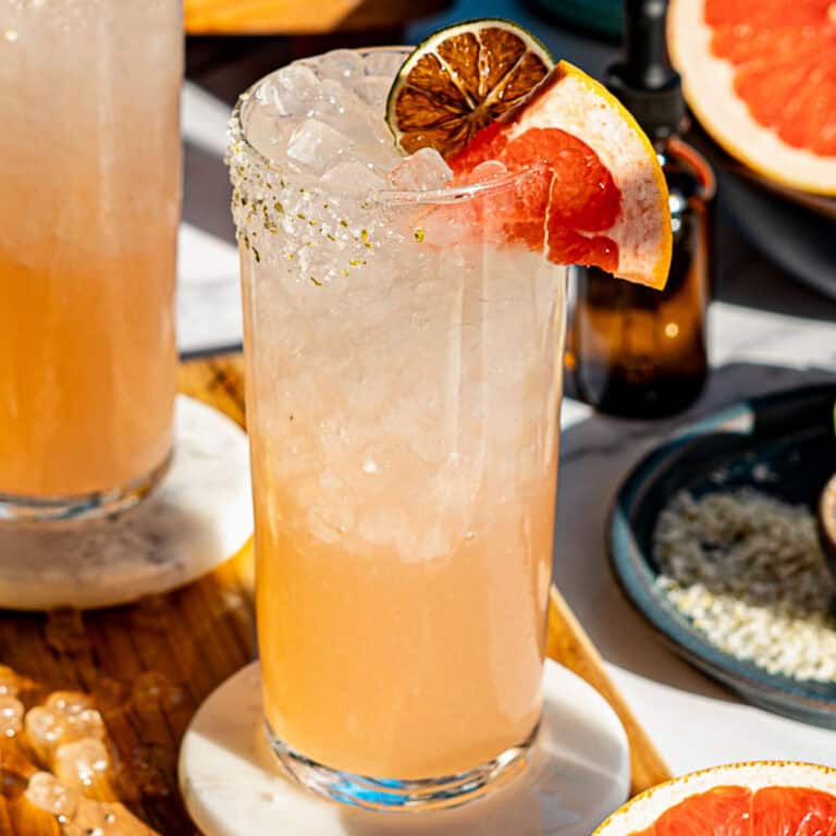 A tall glass filled with a light pink paloma mocktail over crushed ice, garnished with a grapefruit slice and dried lime, sits on a coaster, with another drink and grapefruit pieces nearby.