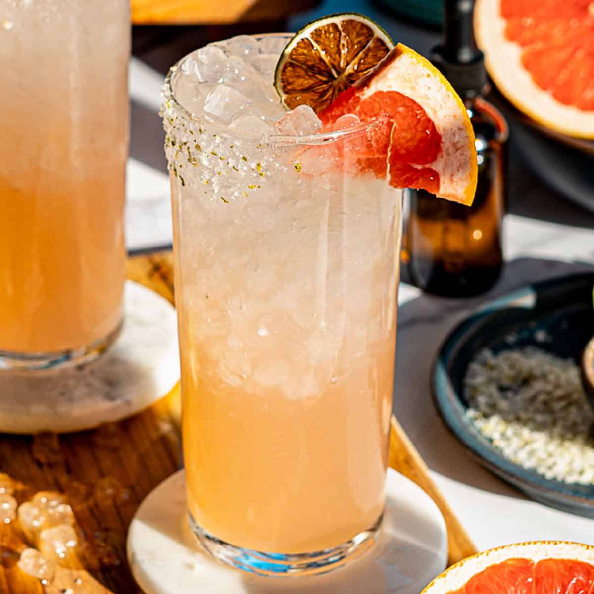 A tall glass filled with a light pink paloma mocktail over crushed ice, garnished with a grapefruit slice and dried lime, sits on a coaster, with another drink and grapefruit pieces nearby.