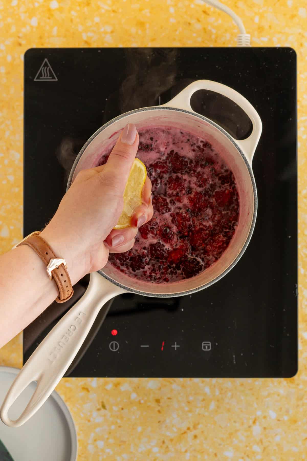 A hand from out of frame is squeezing lemon juice into a saucepan of blackberry simple syrup.