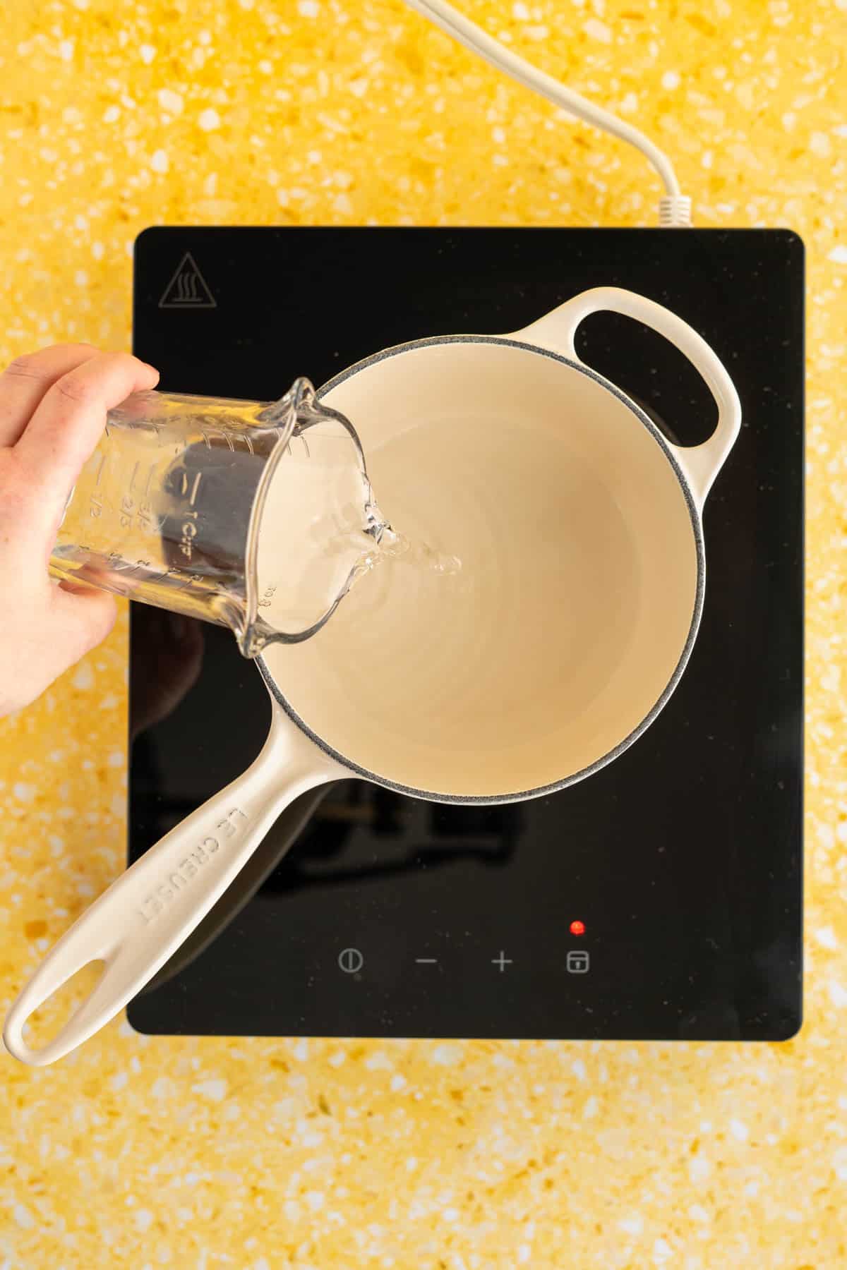 A hand from out of frame is pouring water into a saucepan on an induction heat top.