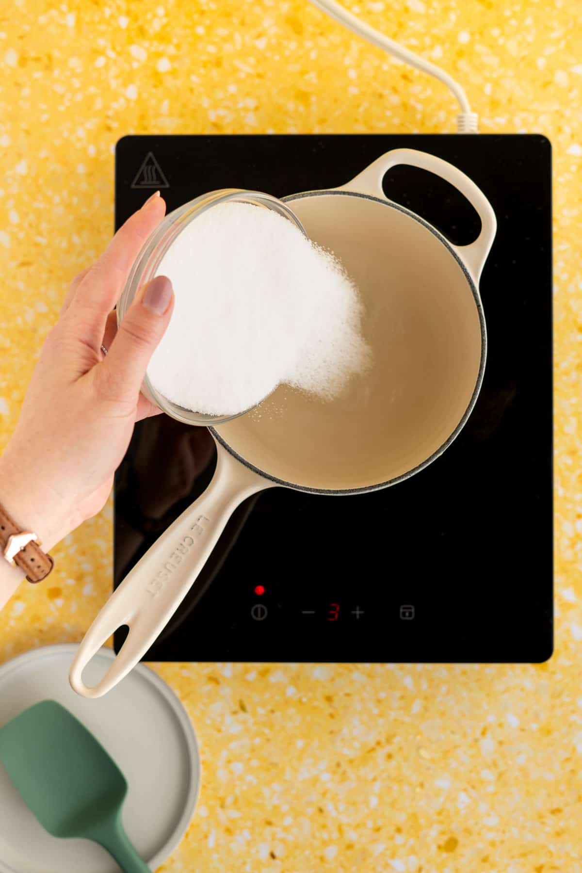 A hand from out of frame is pouring sugar into a saucepan full of water on an induction heat top.