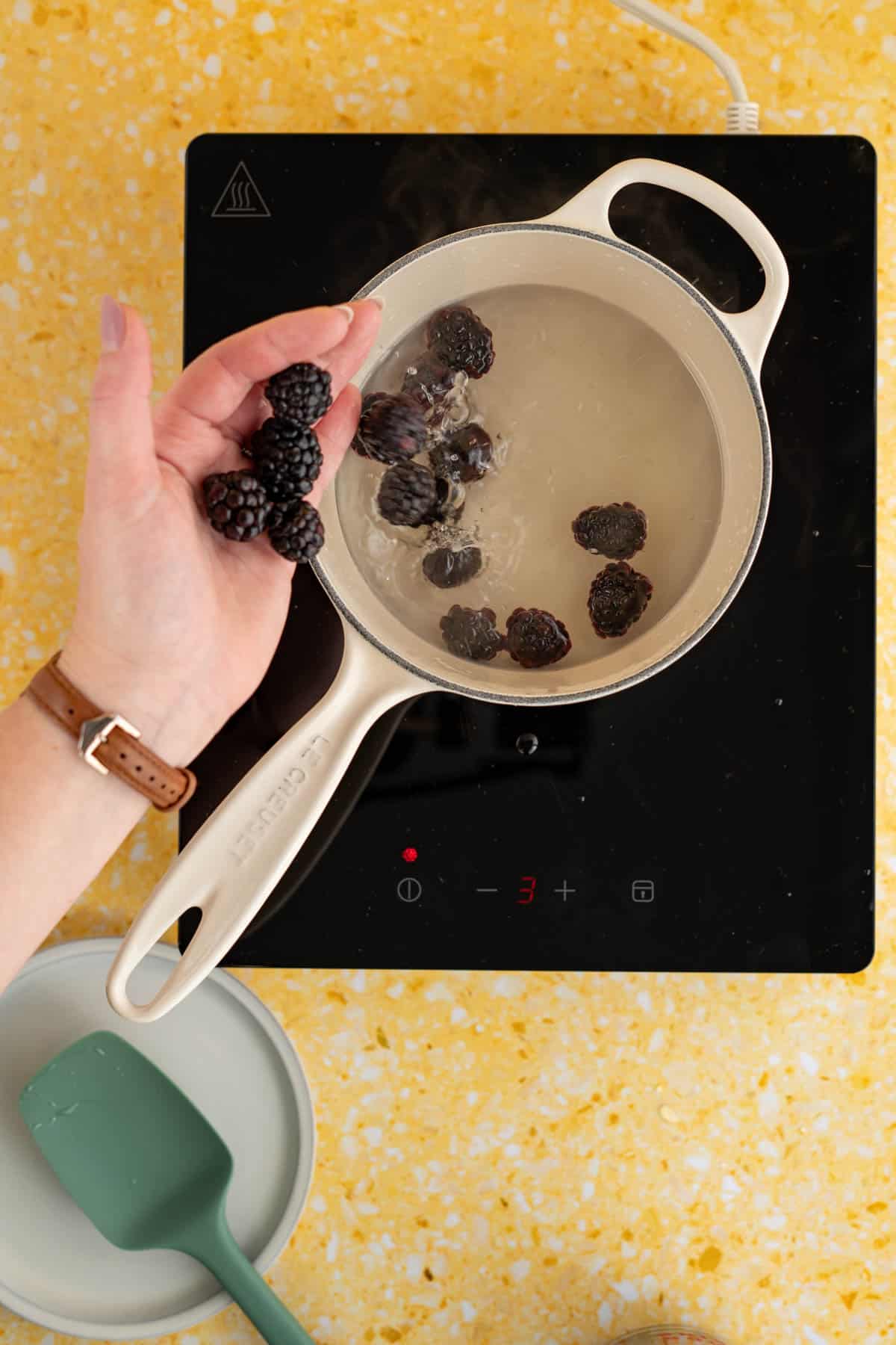 A hand from out of frame is pouring blackberries into a saucepan of simple syrup.
