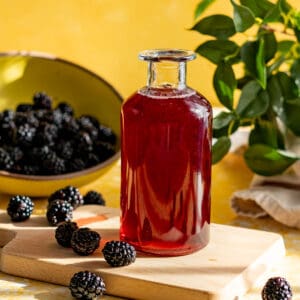 A bottle of homemade blackberry syrup is on a small cutting board with a bowl of blackberries in the background. There are blackberries scattered on the countertop.