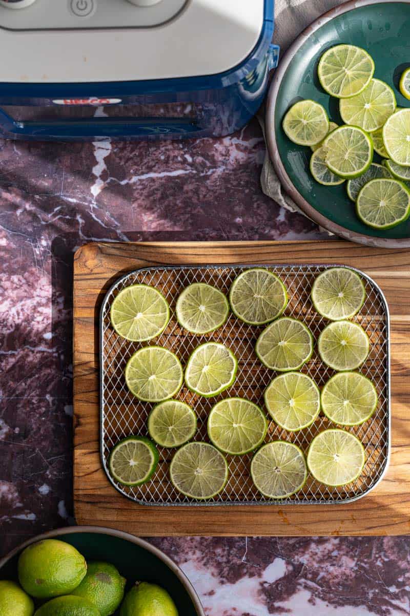 A dehydrator tray that has been filled with limes sits on a wooden cutting board on a marble countertop.