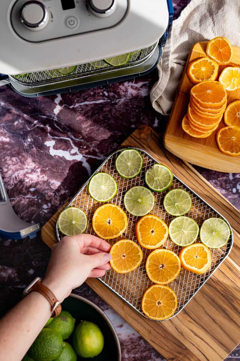 A hand from out of frame is laying sliced of mandarin oranges on a dehydrator tray that has been filled with limes.