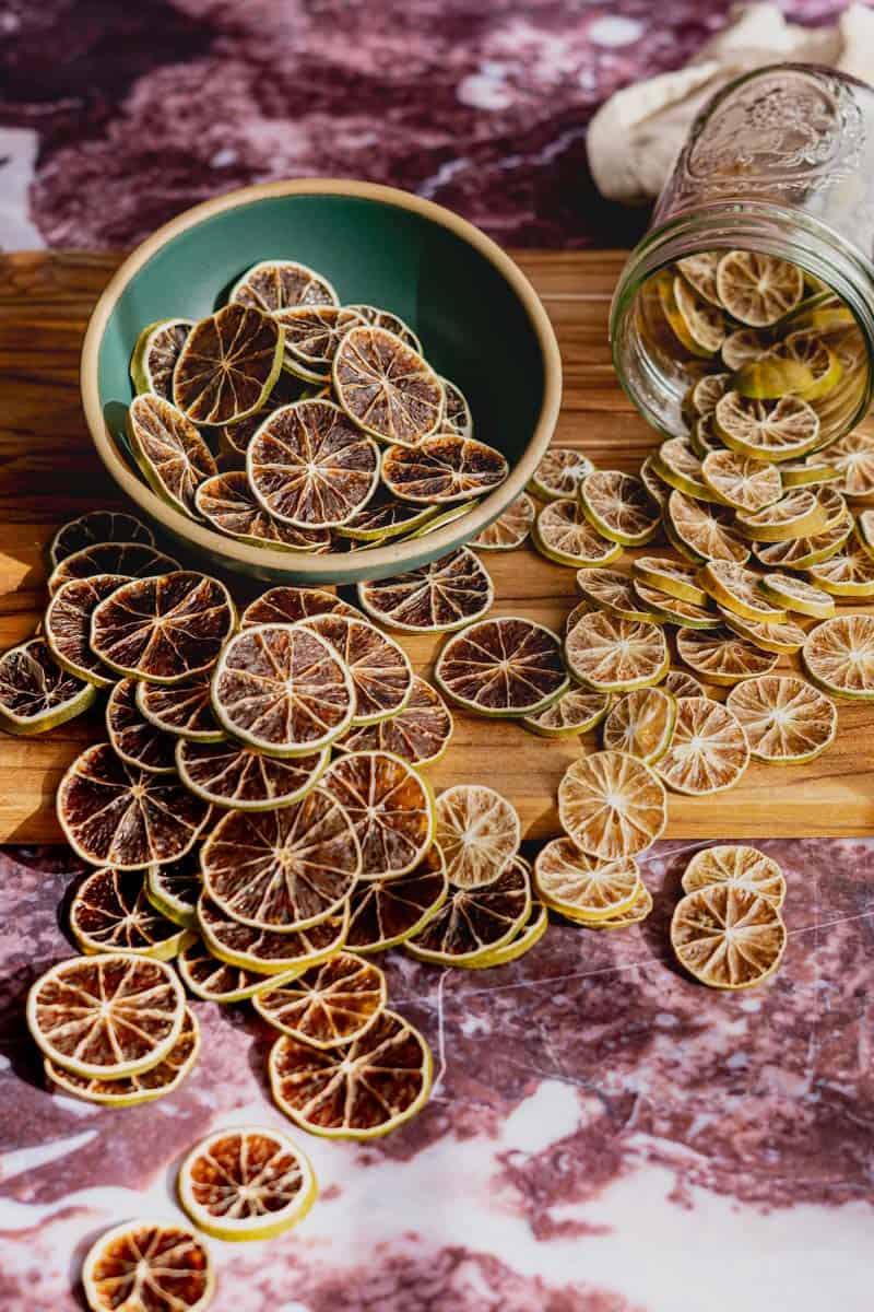 A small bowl of dehydrated limes is spilling onto a wooden cutting board. There are also smaller dehydrated key limes in the frame.