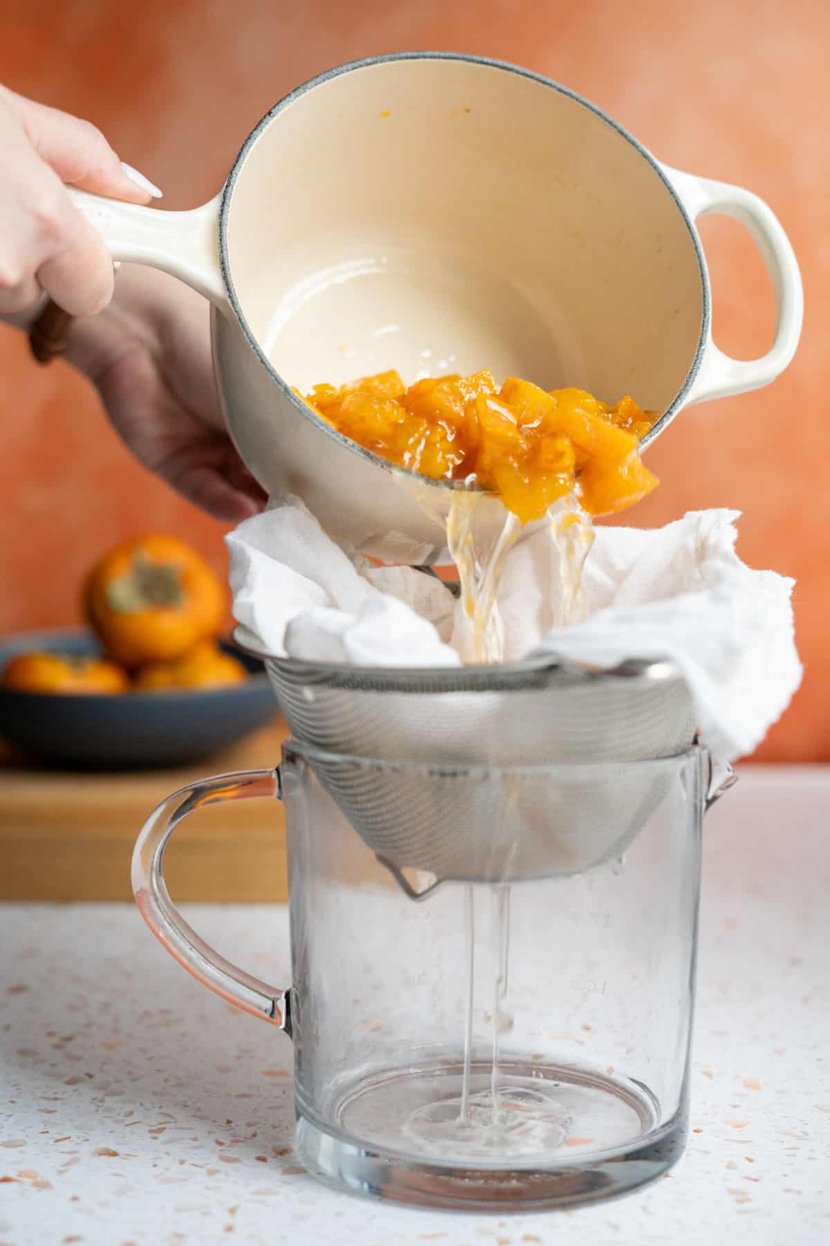 Straining persimmon simple syrup from a saucepan through a fine mesh sieve lined with cheesecloth.