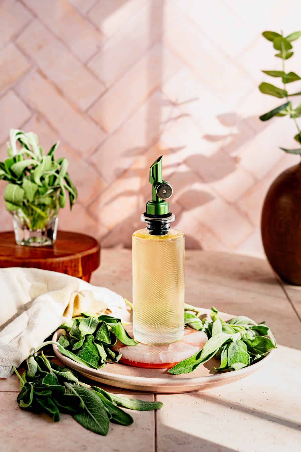 A bottle of sage simple syrup sits on a mauve plate surrounded by sage leaves.
