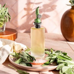 A bottle of sage simple syrup sits on a mauve plate surrounded by sage leaves.