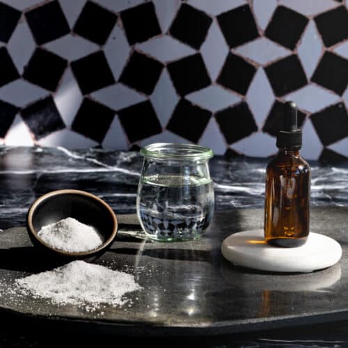 A small bowl of salt and a jar of water sit on a stone countertop next to a dropper bottle of saline solution to use in beverages.