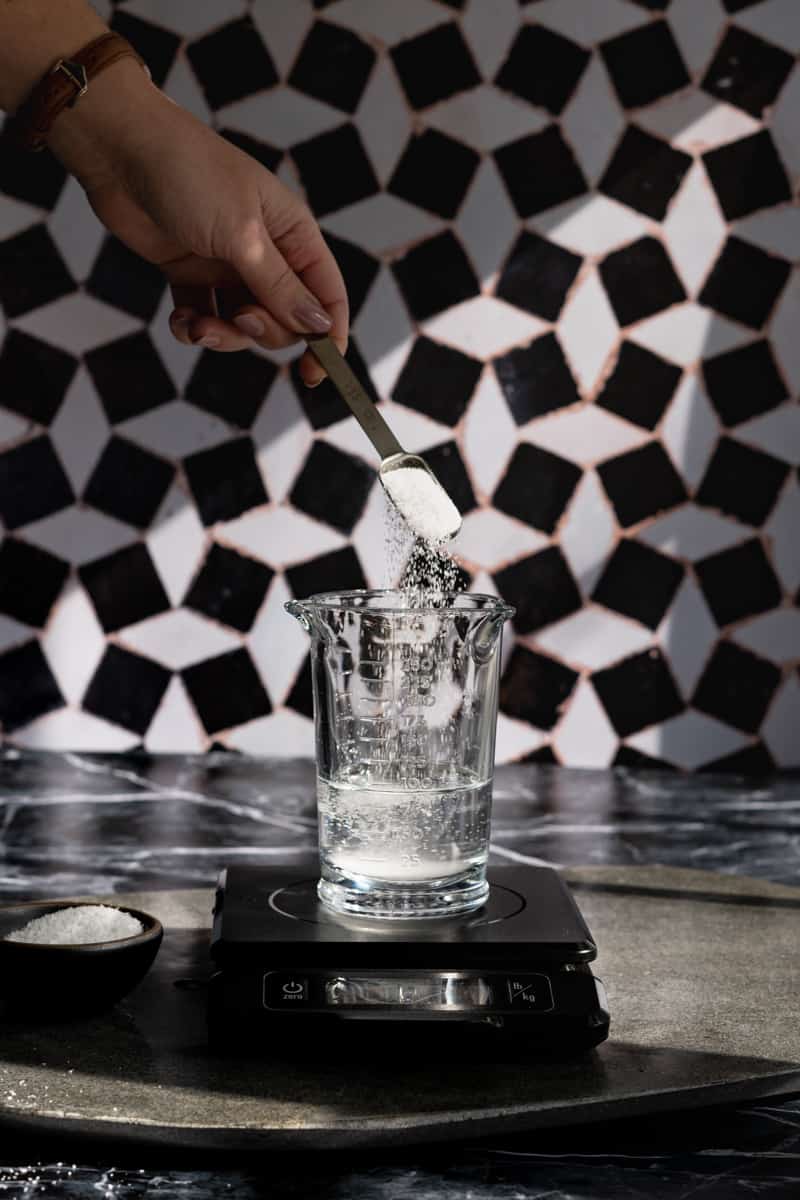 A hand from out of frame adds kosher salt to a measuring glass filled with water.