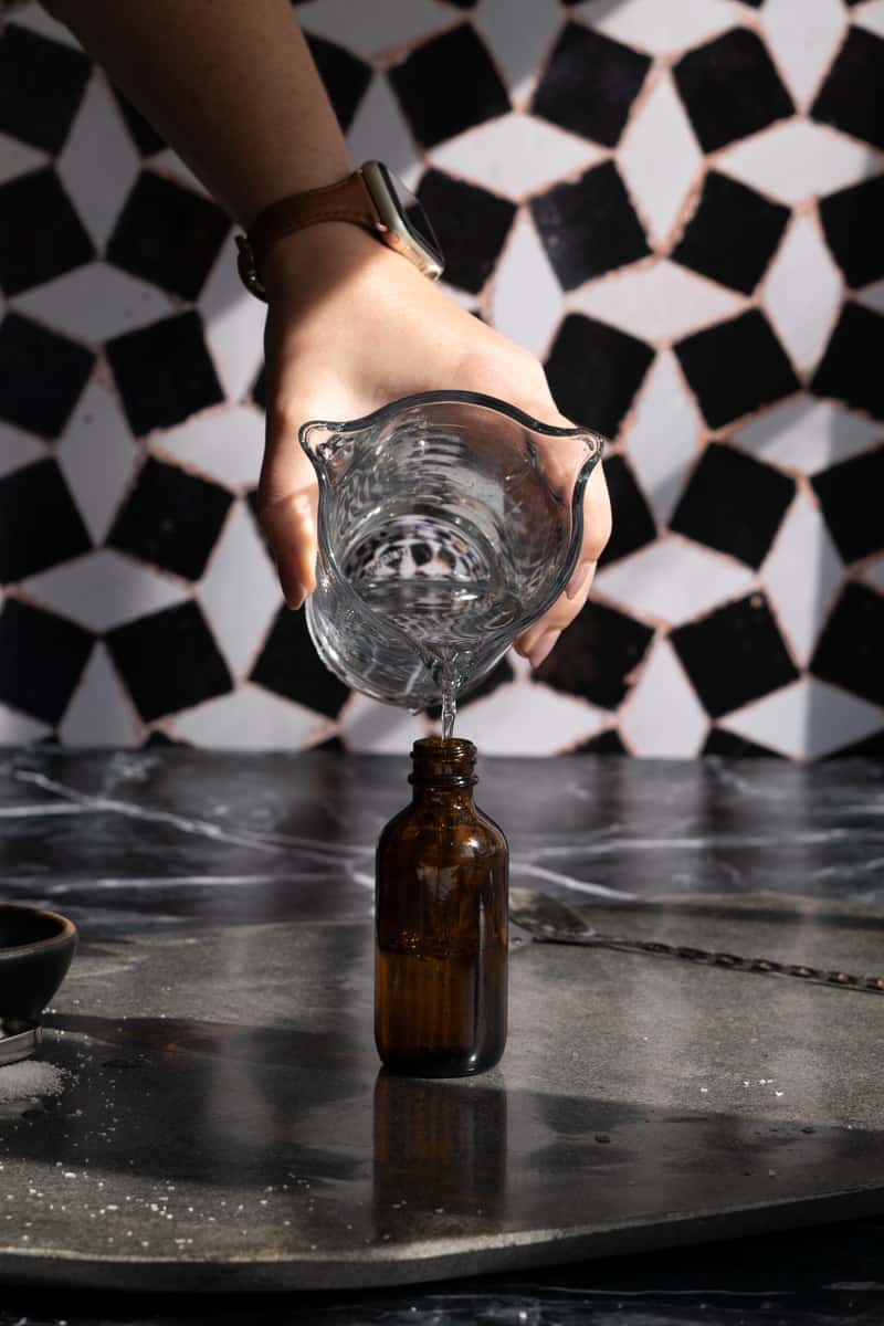 A hand from out of frame is pouring a measuring glass full of saline solution into a dropper bottle for dispensing and storage.