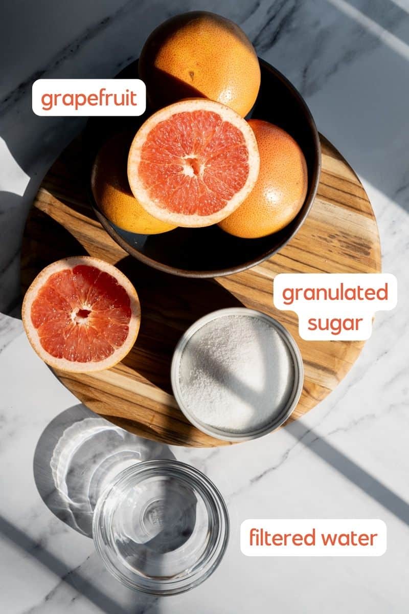 A labeled ingredient shot for a grapefruit syrup recipe. Ingredients used to make the homemade simple syrup sit on a marble countertop on a cutting board.