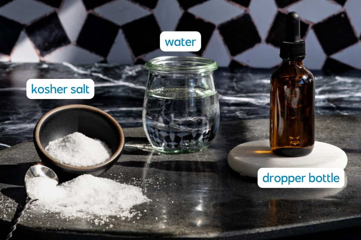 A bowl of kosher salt with a spoon, a glass jar of water, and an amber dropper bottle—perfect for making saline solution for cocktails—are arranged on a dark countertop with a geometric black and white tiled background.