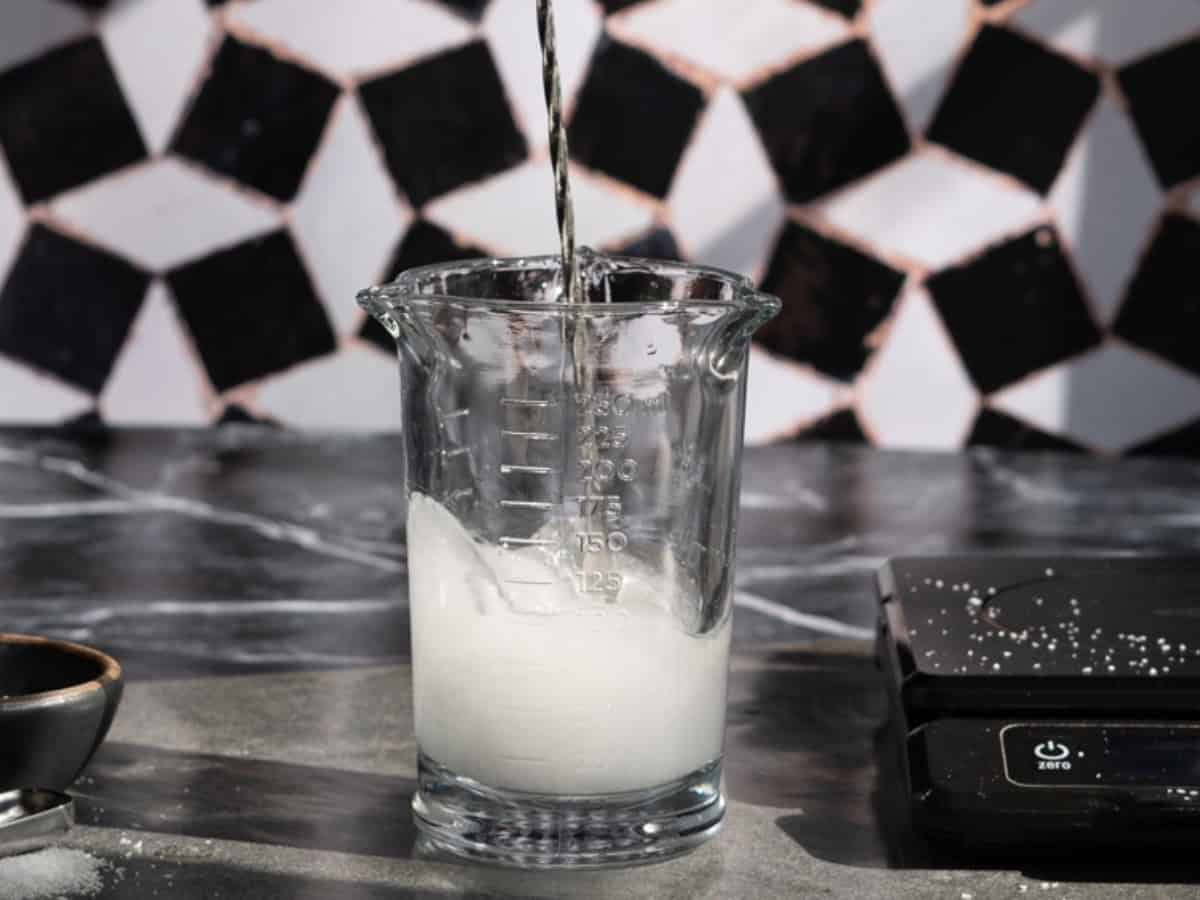 A clear glass measuring cup contains a cloudy white saline solution for cocktails being poured into it. The cup sits on a counter with scattered salt grains, next to a black digital scale and a geometric black-and-white tile wall in the background.